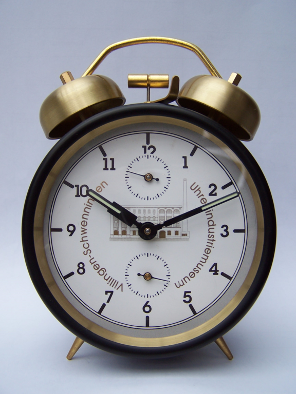 Eine runde, klassische Weckeruhr mit einer weißen Ziffernblatt und schwarzen Ziffern. Die Uhr zeigt zwei Zeiger und einen kleinen Sekundenzeiger. Oben befinden sich zwei Klingeln. Auf dem Ziffernblatt ist eine grafische Darstellung eines Gebäudes abgebildet.